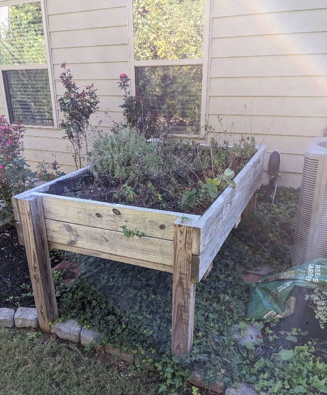 Weathered wooden raised garden bed with overgrown plants beside a house wall.