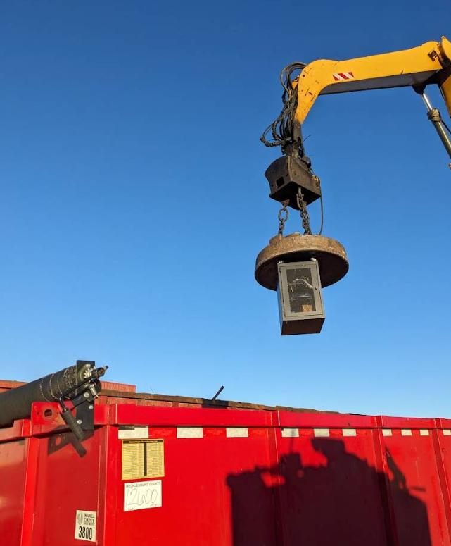 Yellow excavator arm lifts a hanging concrete bucket over a red dumpster under a clear blue sky