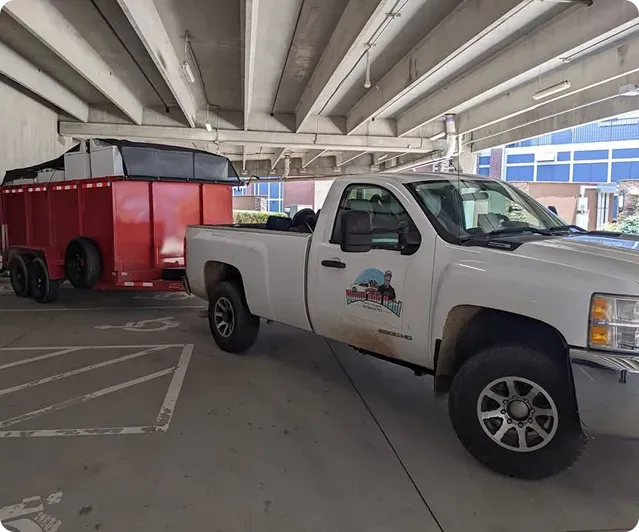A white pickup truck with a logo on its side is parked in a garage, towing a large red dump trailer.