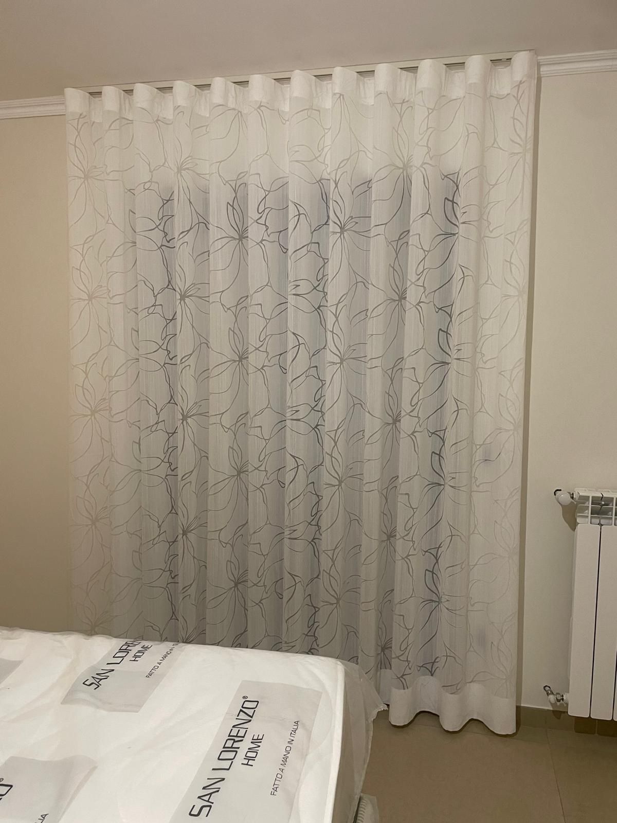 Una tenda bianca trasparente con linee grigio scuro è appesa sopra una finestra; in primo piano c'è un letto con un piumone bianco.