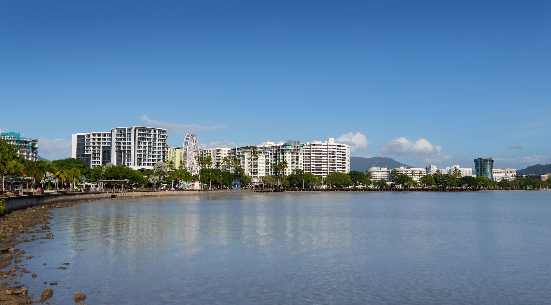 Waterfront view of the Cairns Esplanade skyline featuring high-rise hotels, apartment buildings, and a large Ferris wheel reflecting on the calm water of Trinity Inlet in the Cairns city center.