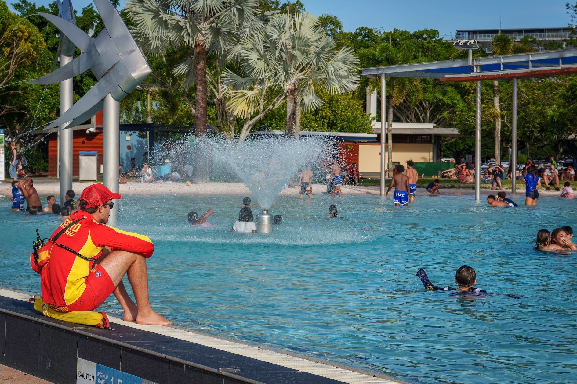 Lifeguard in a red and yellow uniform watching over tourists swimming at the Cairns Esplanade Lagoon public pool near the metal woven fish sculptures in Cairns city center.