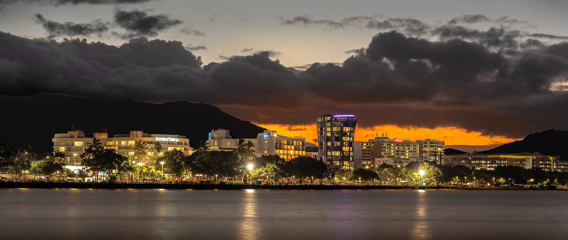 Panoramic view of the Cairns Esplanade waterfront skyline at sunset in Queensland, Australia, featuring illuminated hotels like DoubleTree by Hilton and the Crystalbrook Riley resort tower reflected in Trinity Inlet under a dramatic orange cloud sky.