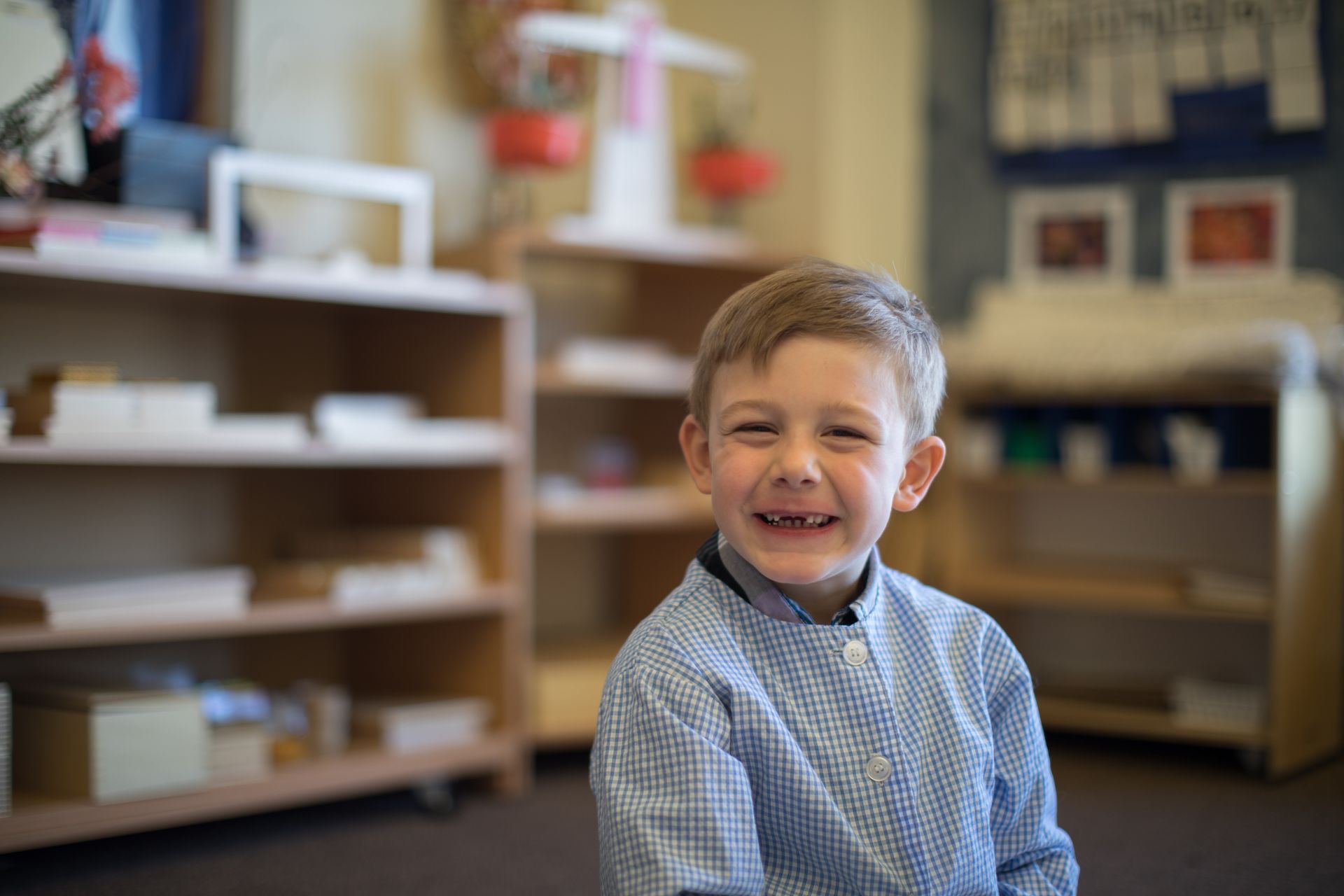 Smiling child sitting in a classroom, with shelves and supplies blurred in the background