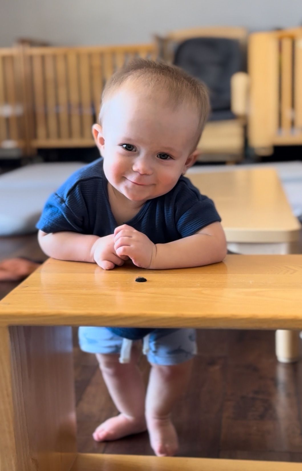 Toddler leaning on a wooden table, smiling indoors in a room with a crib in the background