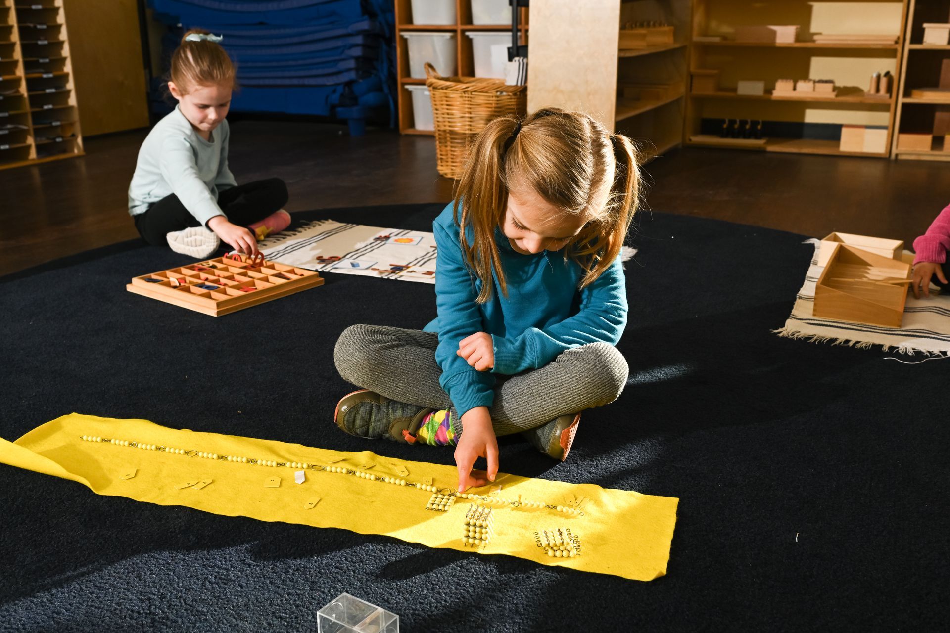 Children playing with blocks on a classroom floor, one on a yellow strip, another by wooden tiles