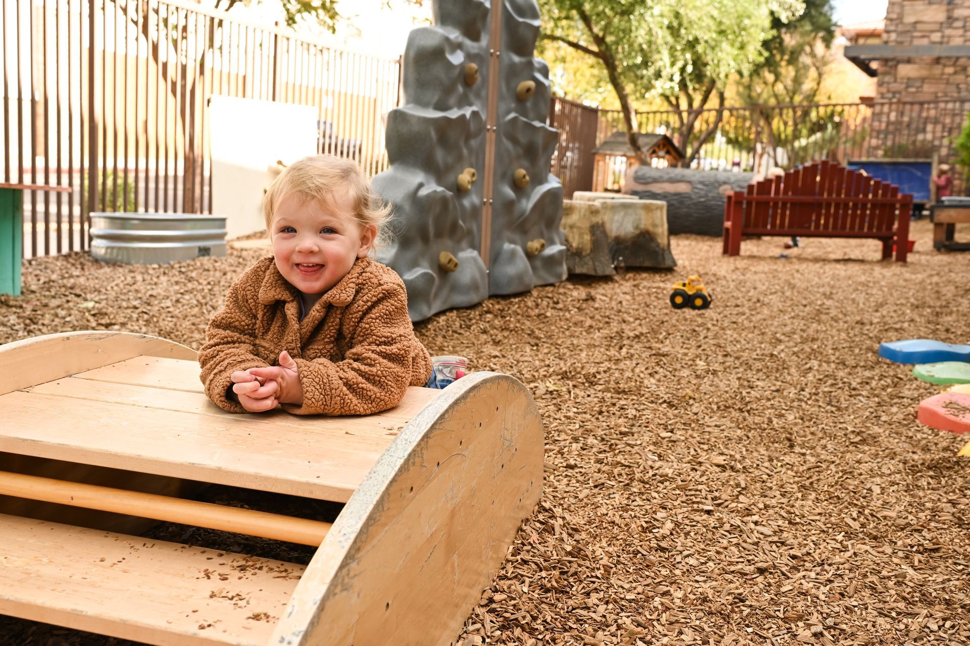 Child leaning on a wooden playground log, smiling in a woodchip play area with climbing rocks and trees.