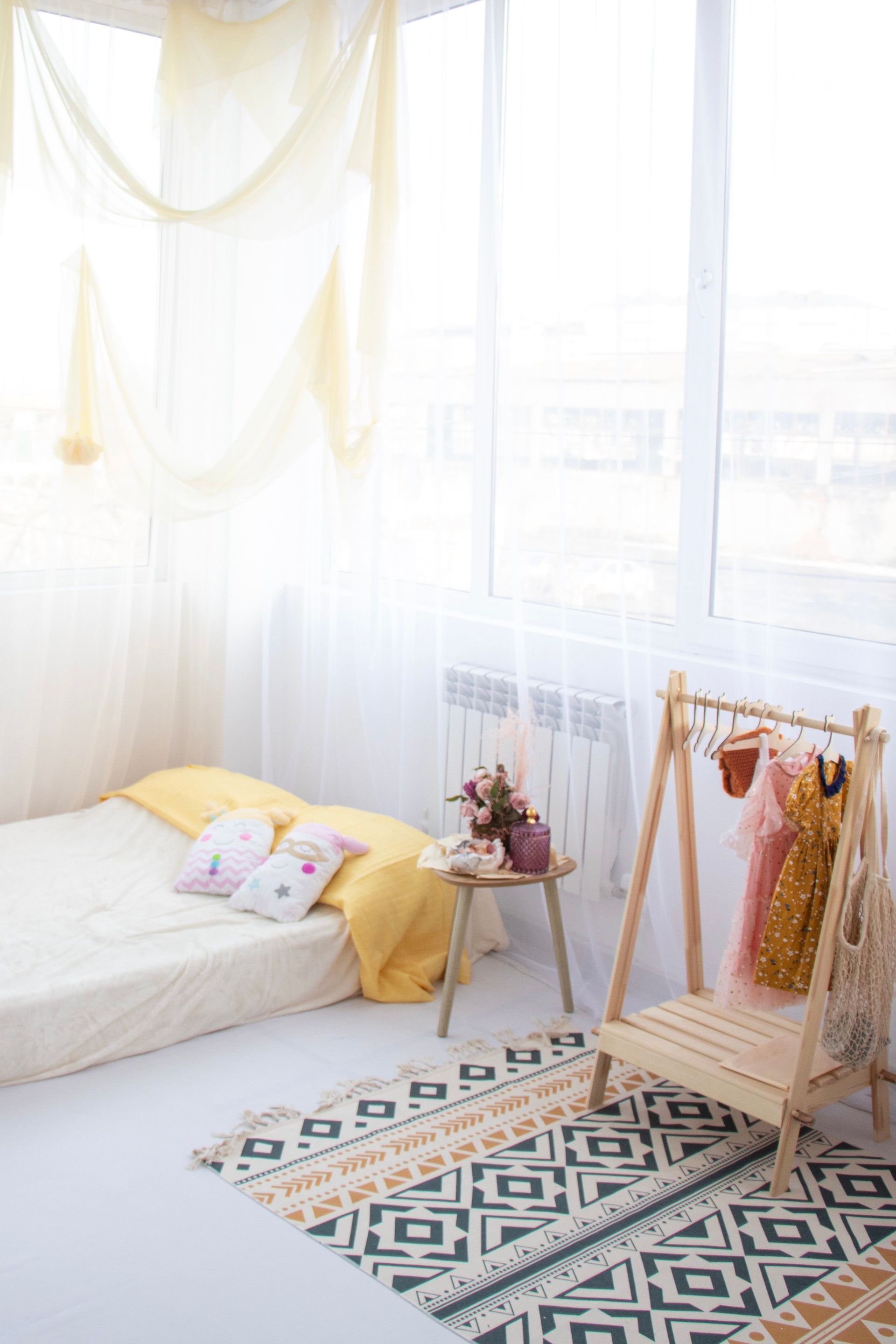 Bright airy bedroom with a low bed, patterned rug, and wooden clothing rack by a sunlit window