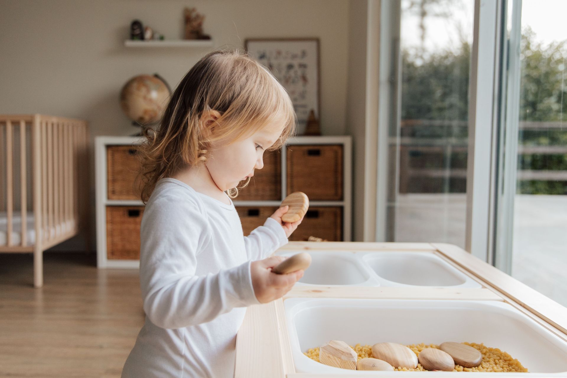 Child standing at a highchair, eating from a tray in a bright kitchen.
