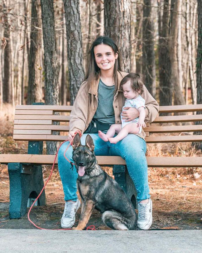 Woman sitting on park bench, holding a baby, with a German Shepherd dog. Outdoor setting with trees.