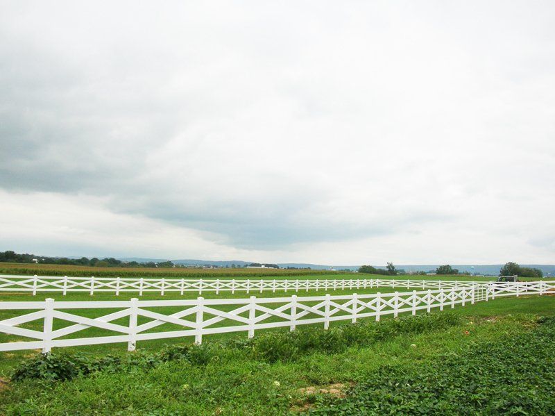 White fence curving through a green field under a cloudy sky, with hills in the distance