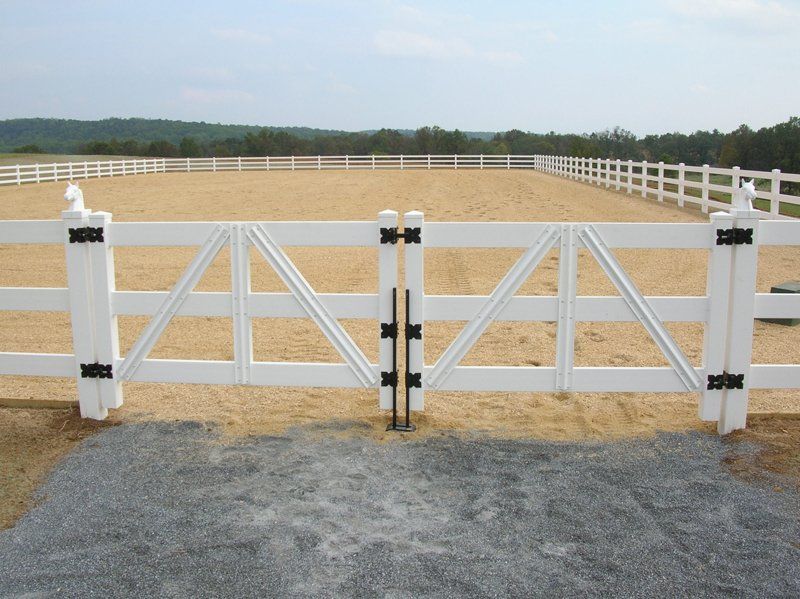 White wooden farm gate with black latches across a gravel driveway, fenced pasture behind it.