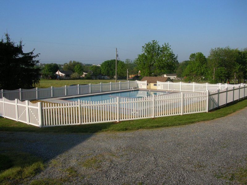 White fenced swimming pool in a grassy yard with gravel foreground under a clear sky
