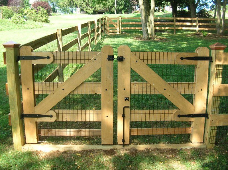 Wooden farm gate with diagonal braces in a sunny grassy yard, backed by a fenced pasture and trees