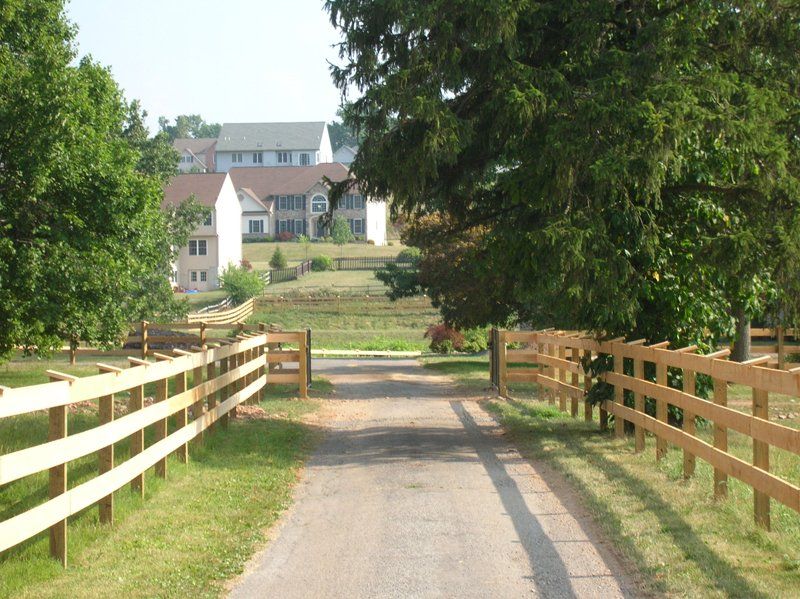 Gravel driveway lined with wooden fences leading to a large house behind trees