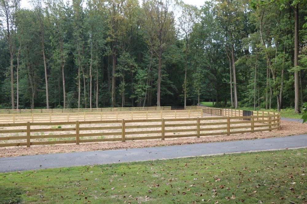 Wooden fence beside a paved path in a grassy field, with tall trees in the background.