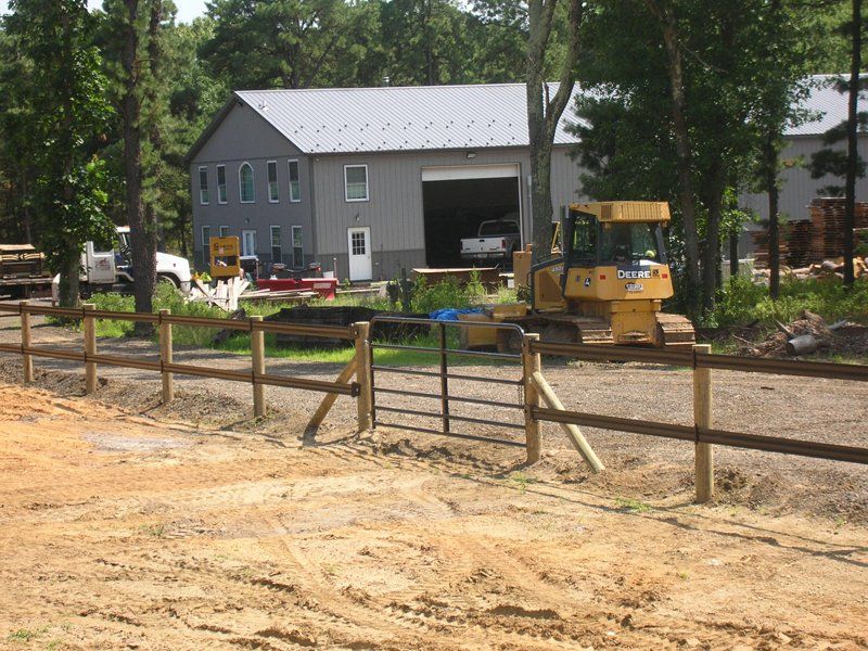 Construction site with yellow bulldozer near a gray barn and wooden fence in a wooded area