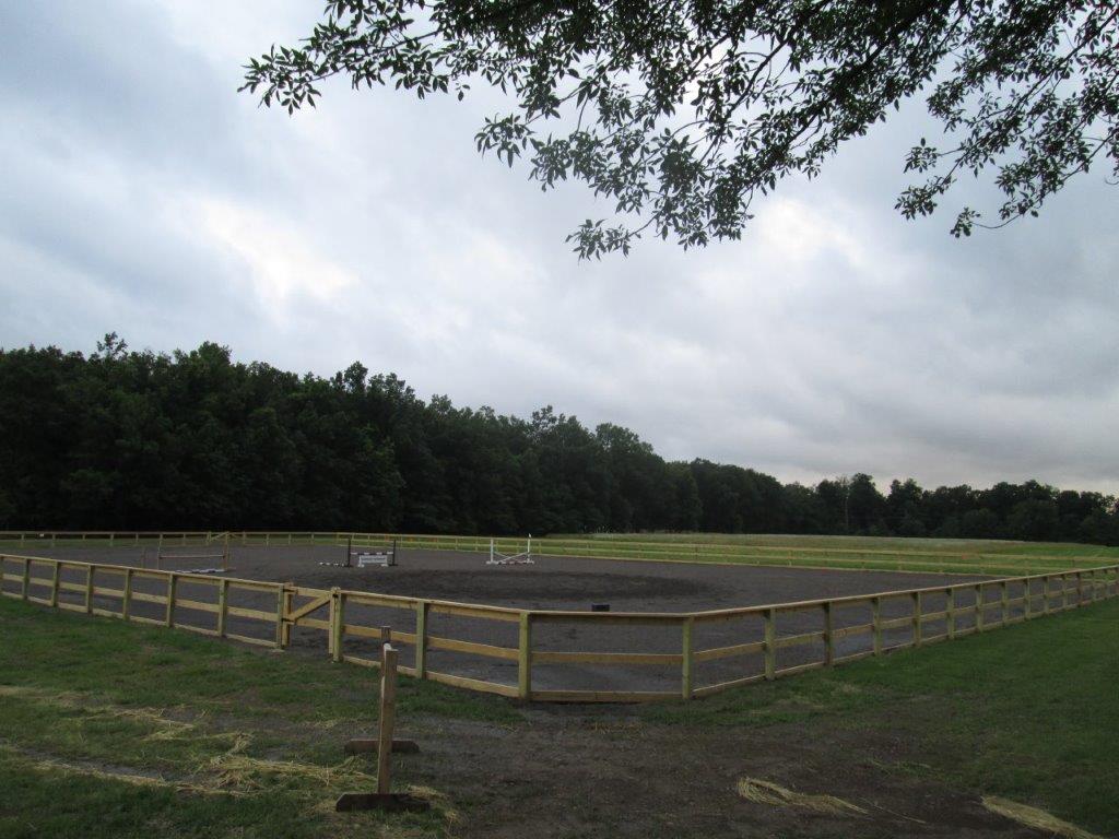 Open dirt riding ring with yellow fence, surrounded by trees under an overcast sky