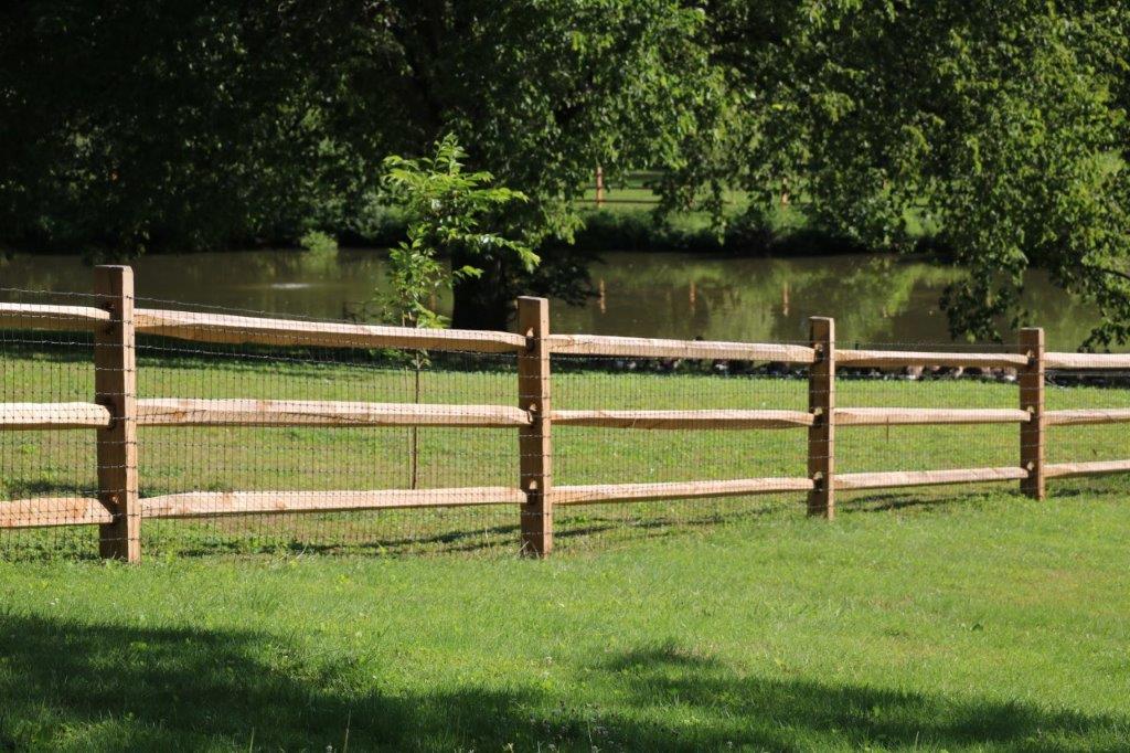 Wooden fence beside a grassy field with trees and a pond in the background