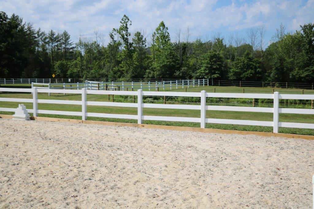 White fence along a gravel path beside a grassy field and trees under a blue sky
