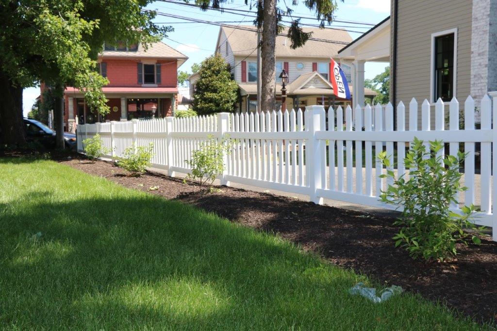 White picket fence along a grassy yard with trees and houses in the background