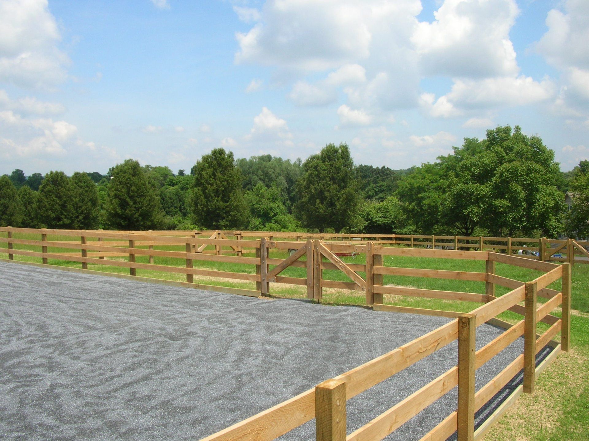 Wooden fence around a gravel paddock with green trees and a partly cloudy sky