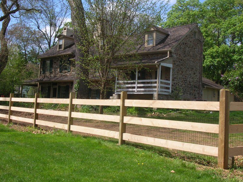 Stone farmhouse behind a wooden fence, with trees and green lawn in the foreground.