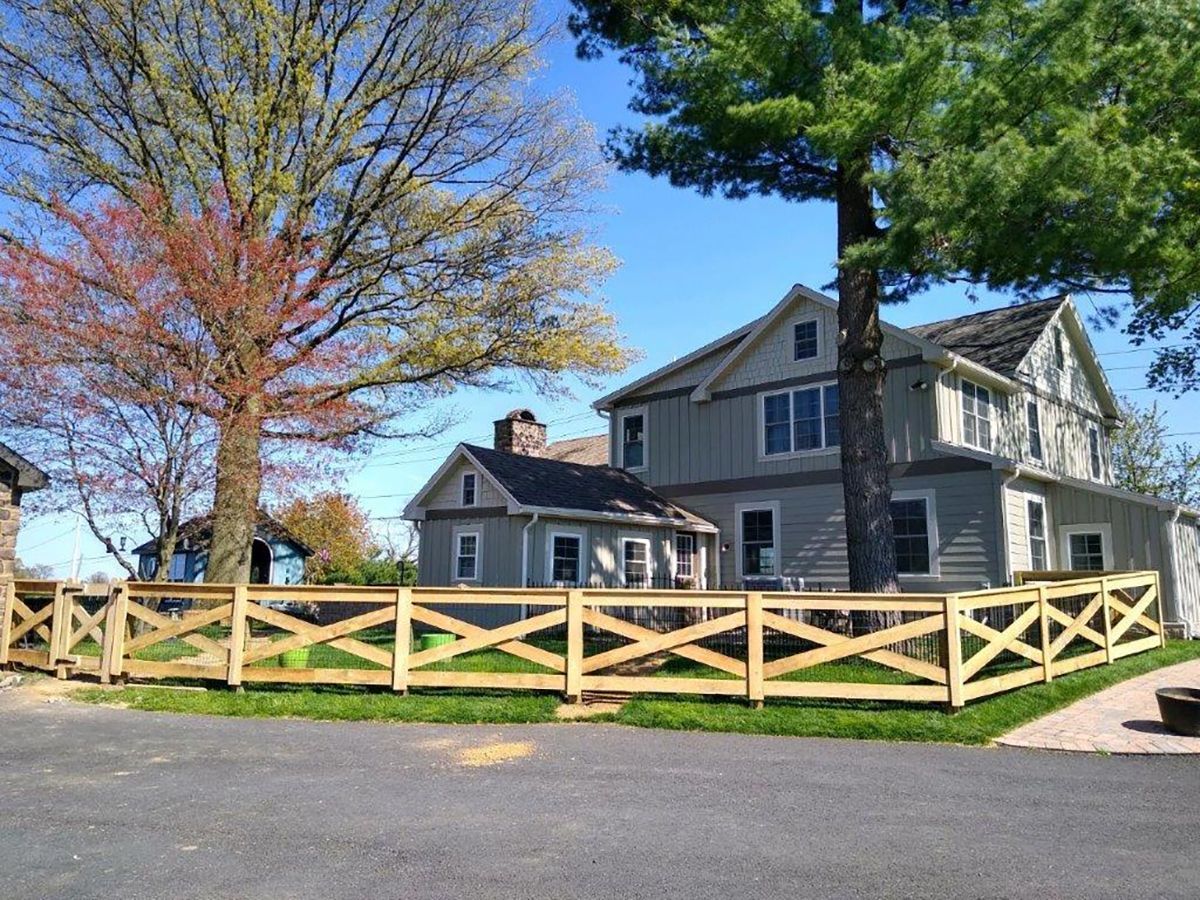 Gray house with a wooden fence, two large trees, and a driveway under a bright blue sky