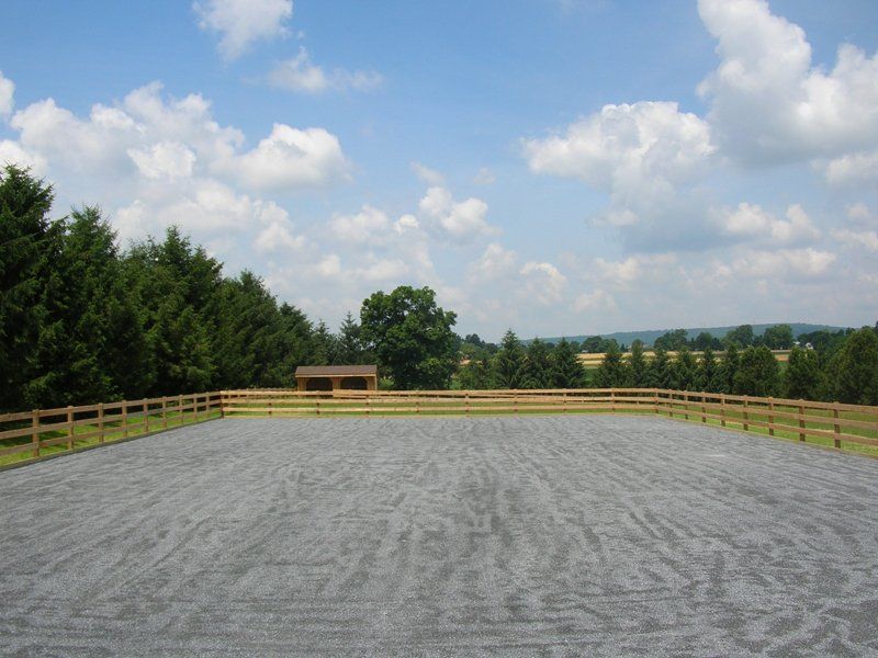 Gravel riding arena with wooden fence under a partly cloudy sky, surrounded by trees and hills