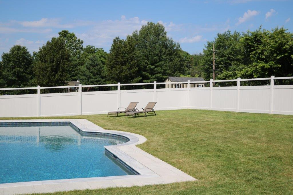 Backyard pool with white fence, green lawn, lounge chairs, and trees under a blue sky