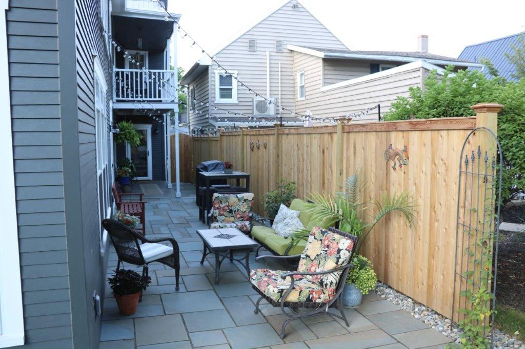 Narrow patio with black chairs, potted plants, and a wooden fence beside apartment buildings