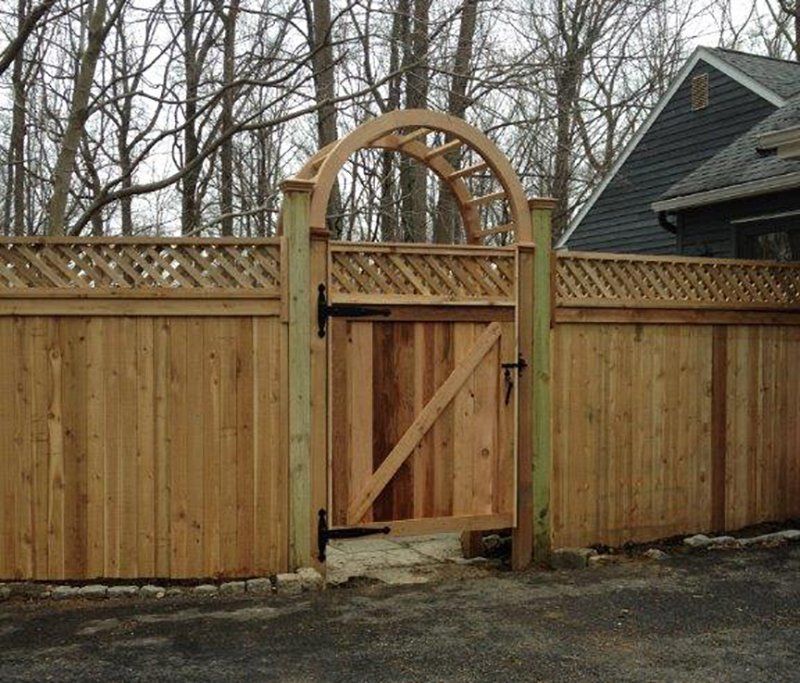 Wooden garden gate with arched trellis in a fenced yard, beside a house and leafless trees