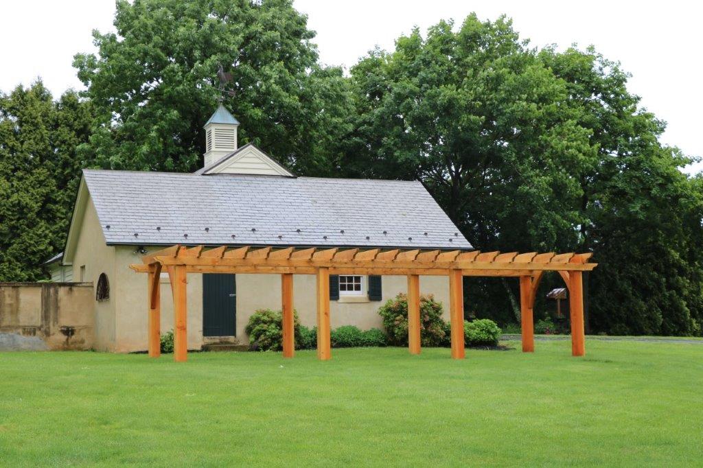Small stone chapel with gray roof beside a wooden pergola on a green lawn, backed by trees