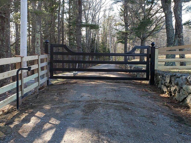 Closed black wooden gate across a gravel driveway in a wooded area