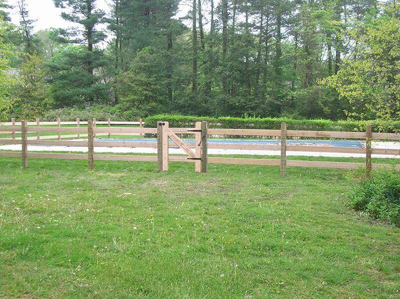 Wooden fence around a grassy yard with trees and a pool in the background.