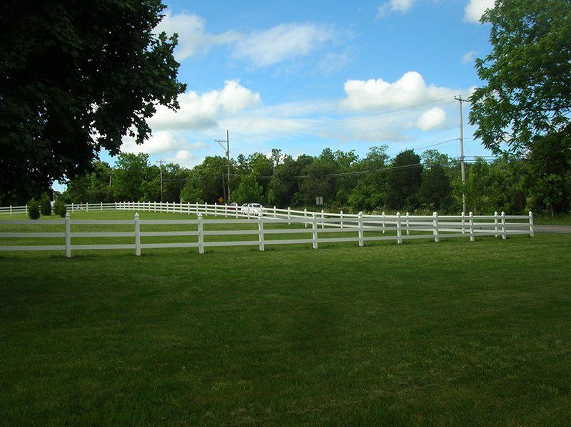 White fence enclosing a green field beneath a blue sky with clouds and trees