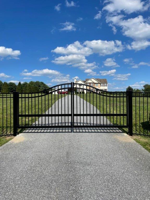 Black metal gate blocking a gravel driveway leading to a large house under a blue sky with clouds