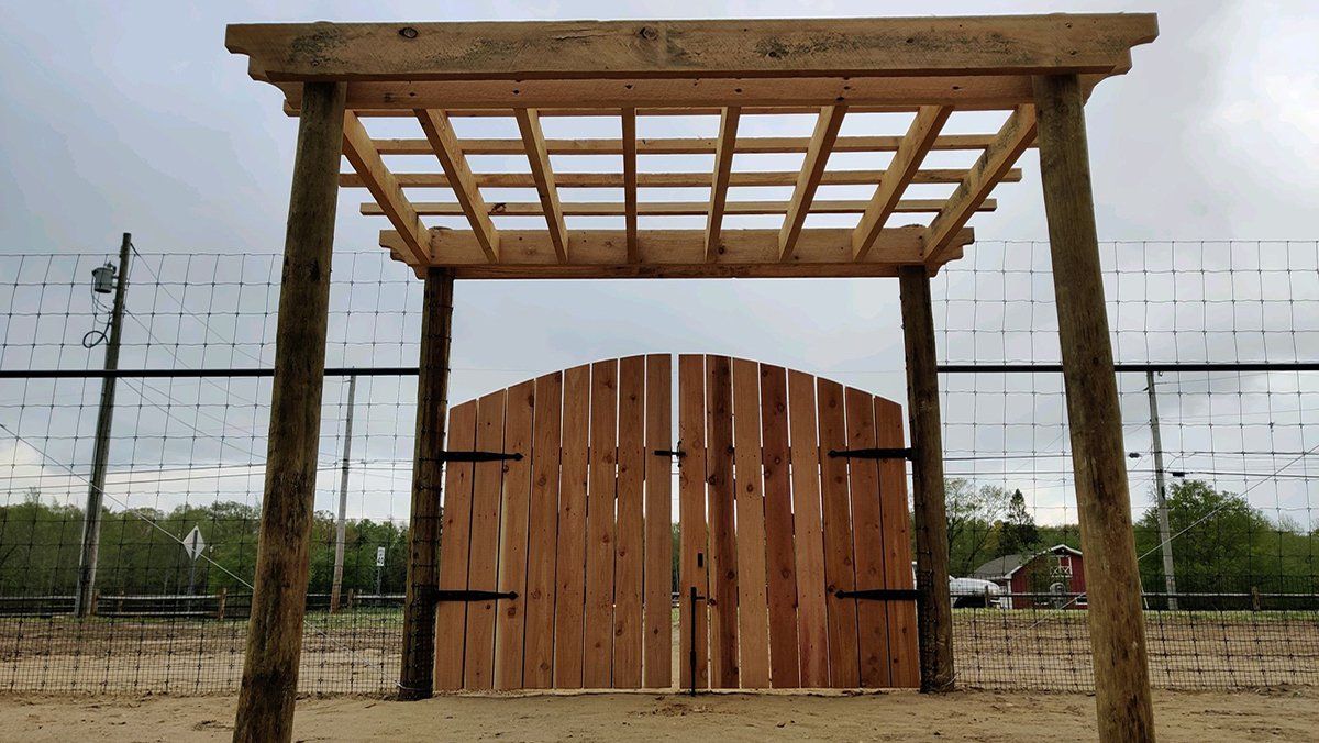 Wooden gate under a simple pergola at a fenced outdoor entrance, with a cloudy sky overhead