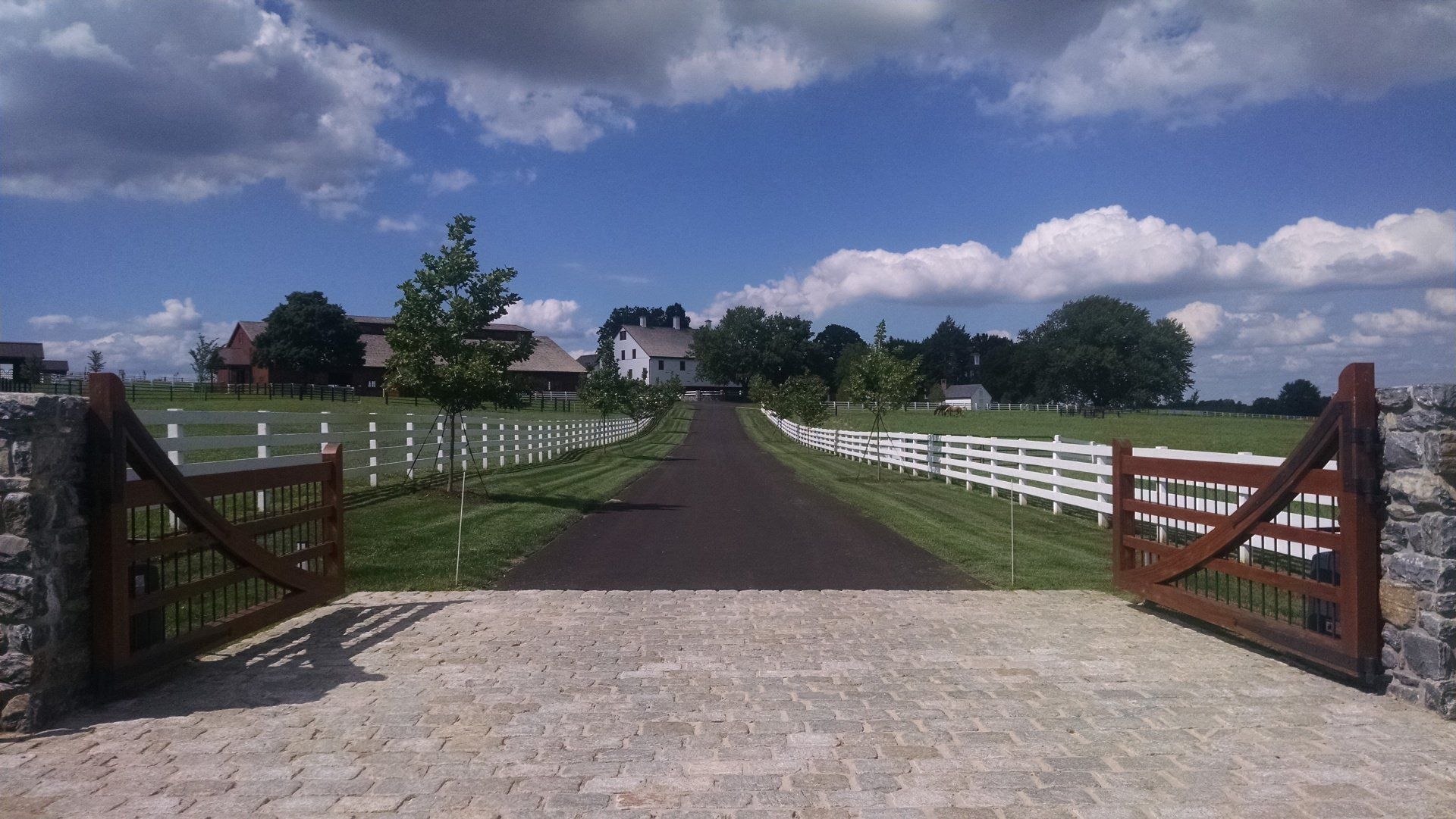 Open white farm gate leading to a paved driveway under a blue sky with clouds