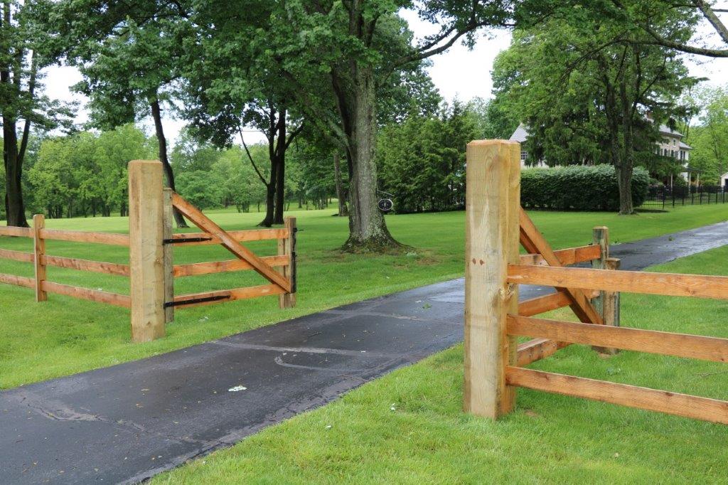 Open wooden gate beside a paved driveway in a green park with trees and grass.