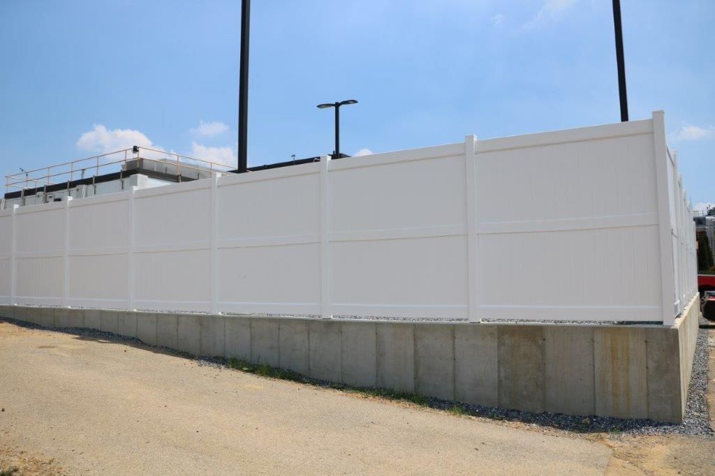 Tall white fence along a roadside under a blue sky, with utility poles and concrete base border
