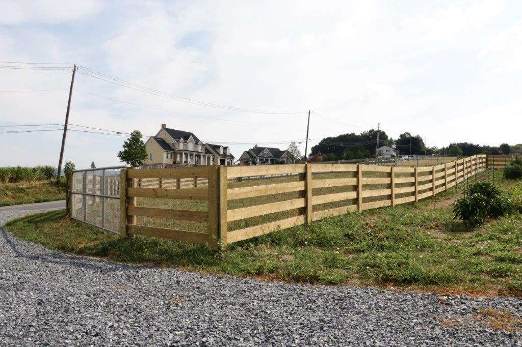 Wooden fenced field beside a gravel road, with a house in the background under a cloudy sky.