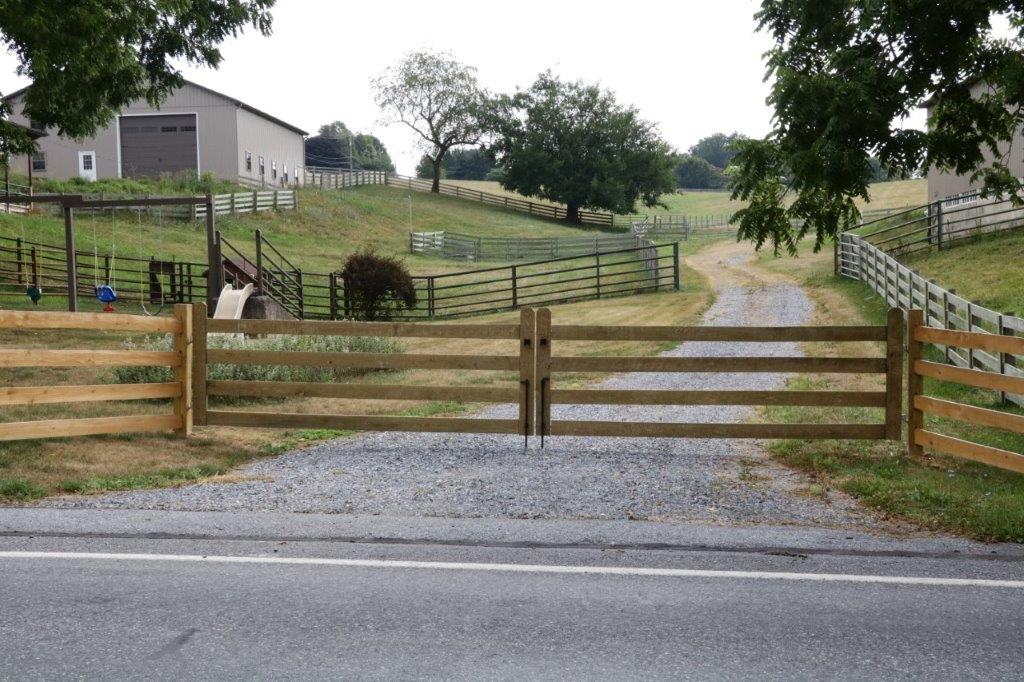 Wooden gate blocking a gravel driveway leading to a farmhouse and fenced pasture.
