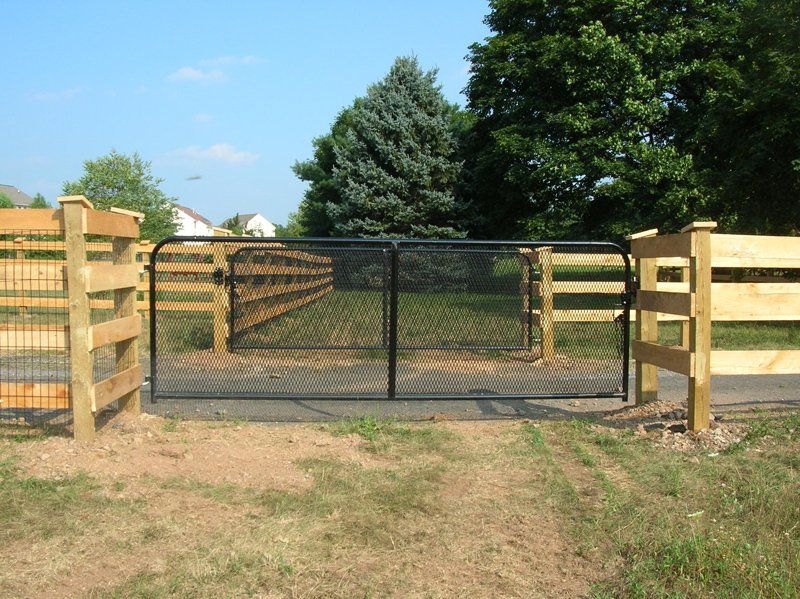 Black metal gate in a wooden fence across a dirt driveway with trees behind it