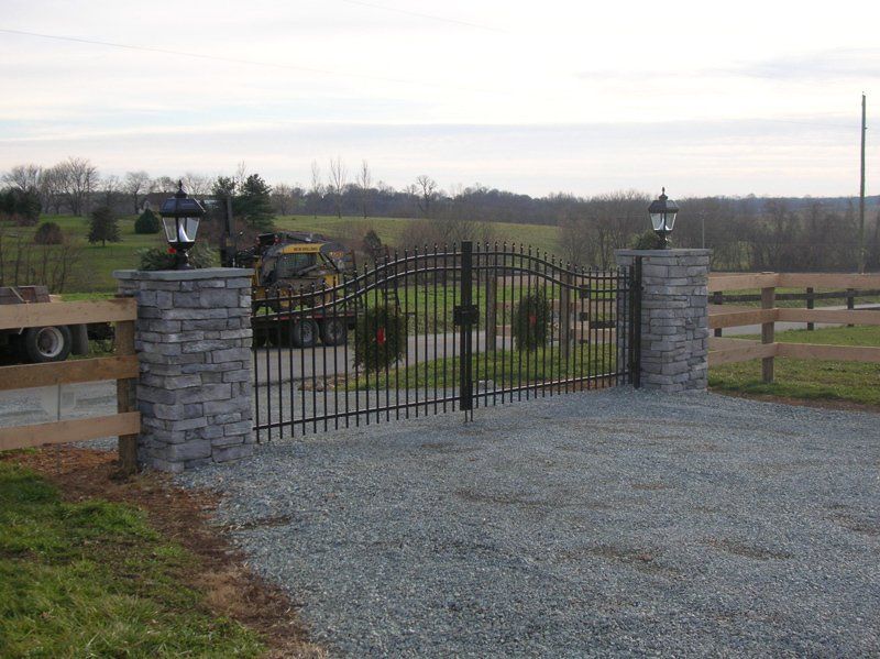 Gravel driveway entrance with black wrought-iron gate between stone pillars and wooden fences in a rural field