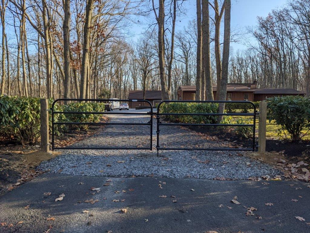 Black metal gate at the end of a gravel driveway, bordered by hedges and bare trees in winter.