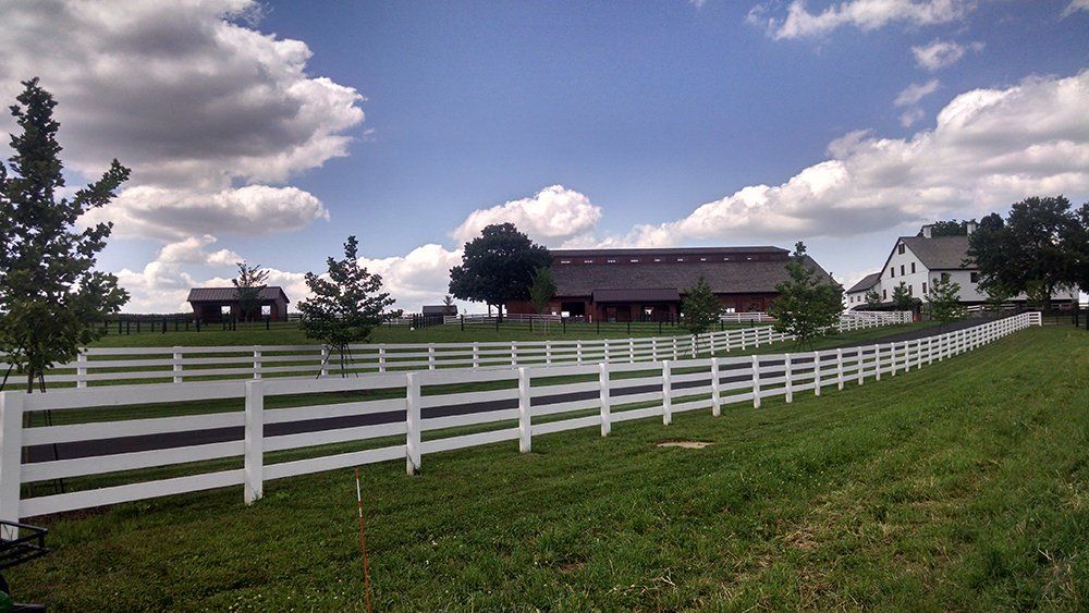 White fence along a green field with barns under a cloudy blue sky
