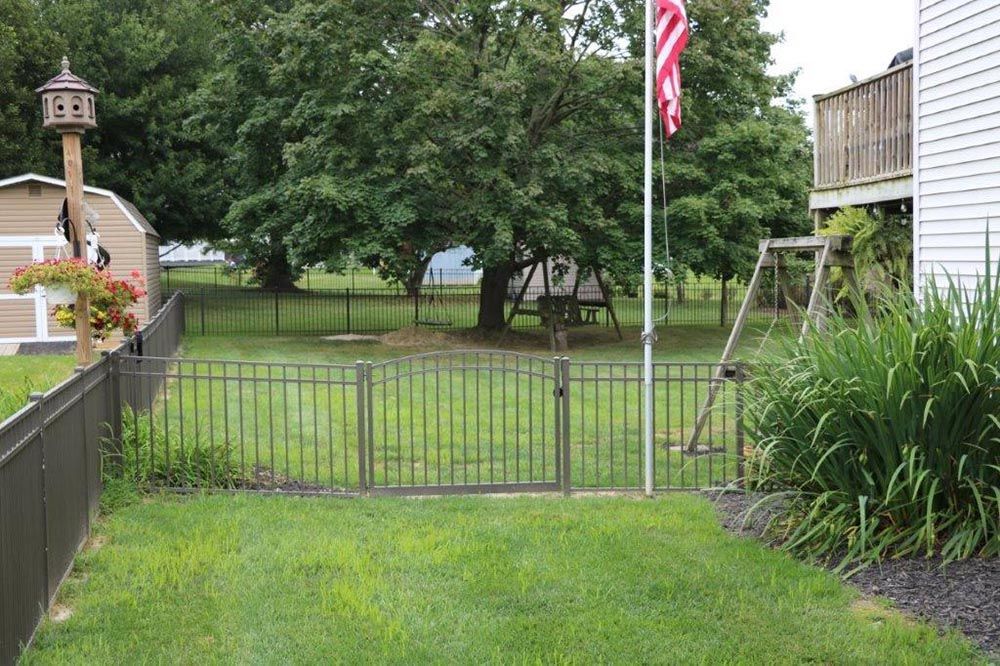 Grassy backyard with a metal gate, fence, swing set, trees, and an American flag by a house