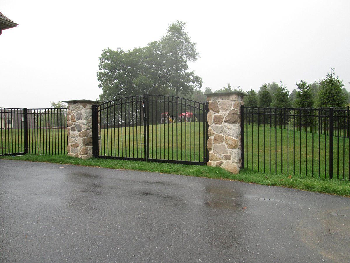 Black metal gate between stone pillars beside a wet driveway and grassy yard.