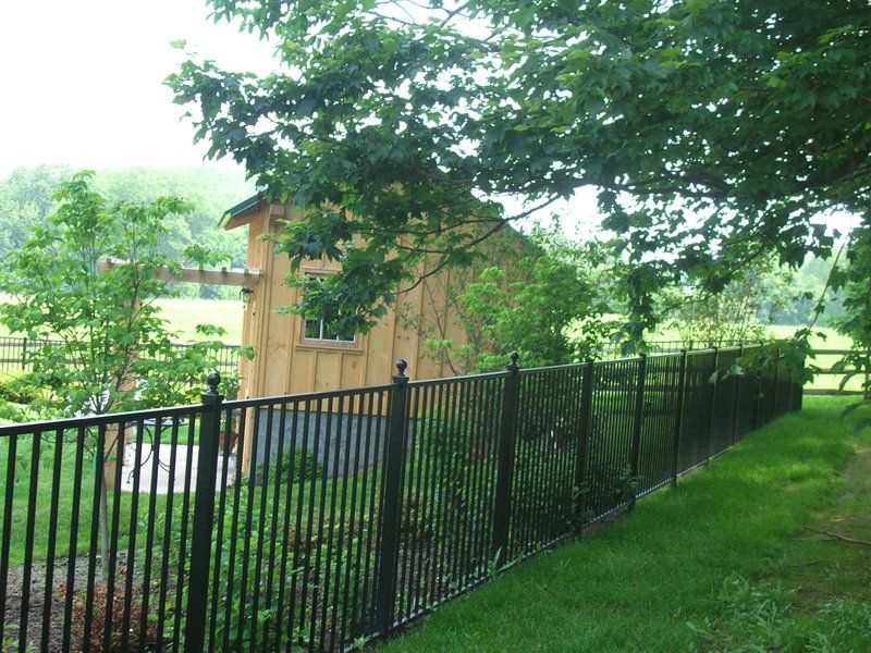 Black metal fence along a grassy slope beside a tan building and leafy trees.